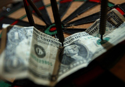 Photograph of darts pinning a dollar bill to a dart board.