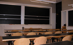 A classroom with three long tables spanning the classroom, chairs behind each table, and two sliding chalkboards at the front.