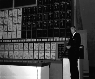 Black and white photo of Glenn T. Seaborg in front of the periodic table of elements. Black and white photo of Glenn T. Seaborg in front of the periodic table of elements.