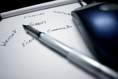 Photograph of computer mouse, and pen laying on a pad of paper that has the word &quot;writing&quot; on it in numerous languages.