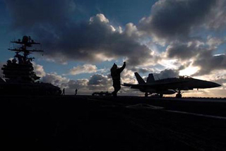 A photo of a silhouetted F-18C Hornet landing on an aircraft carrier.