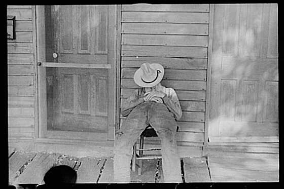 Photo of a man with a hat over his face slumped in a chair on a porch.